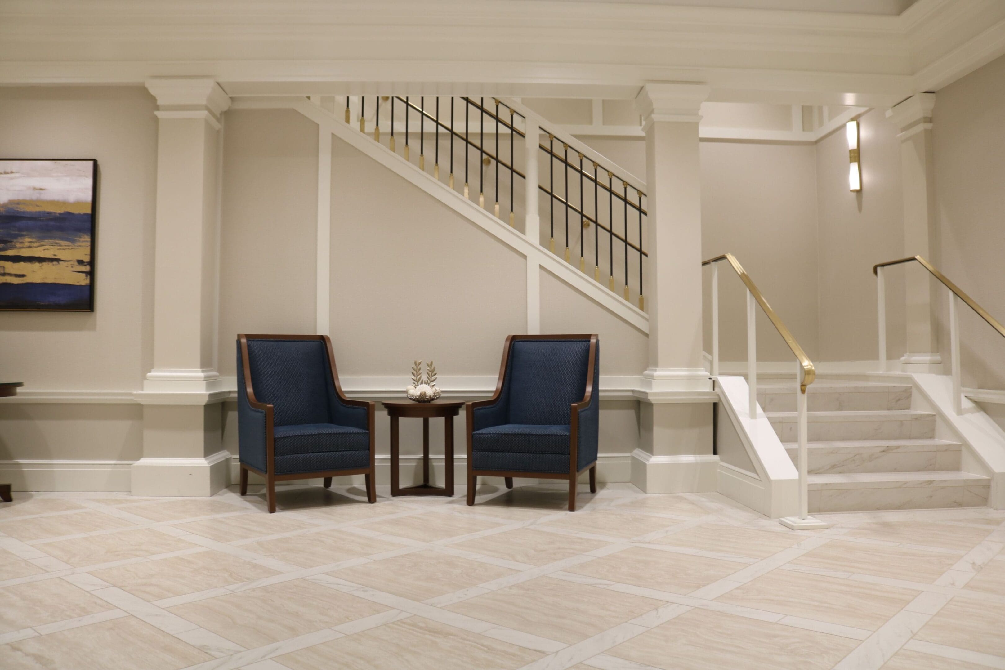 Stylish lobby with two blue chairs and a small table in front of a white staircase. Neutral tones, tiled floor, and a modern art piece on the wall.