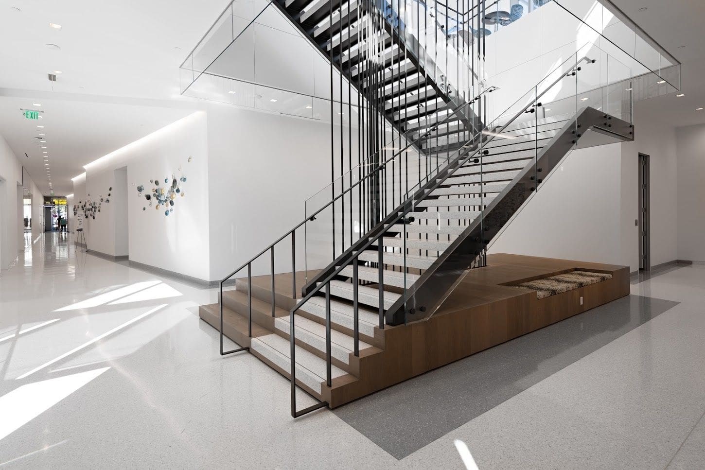 Modern interior with a floating glass and metal staircase, white walls, and terrazzo flooring. Decorative wall art is visible in the hallway.