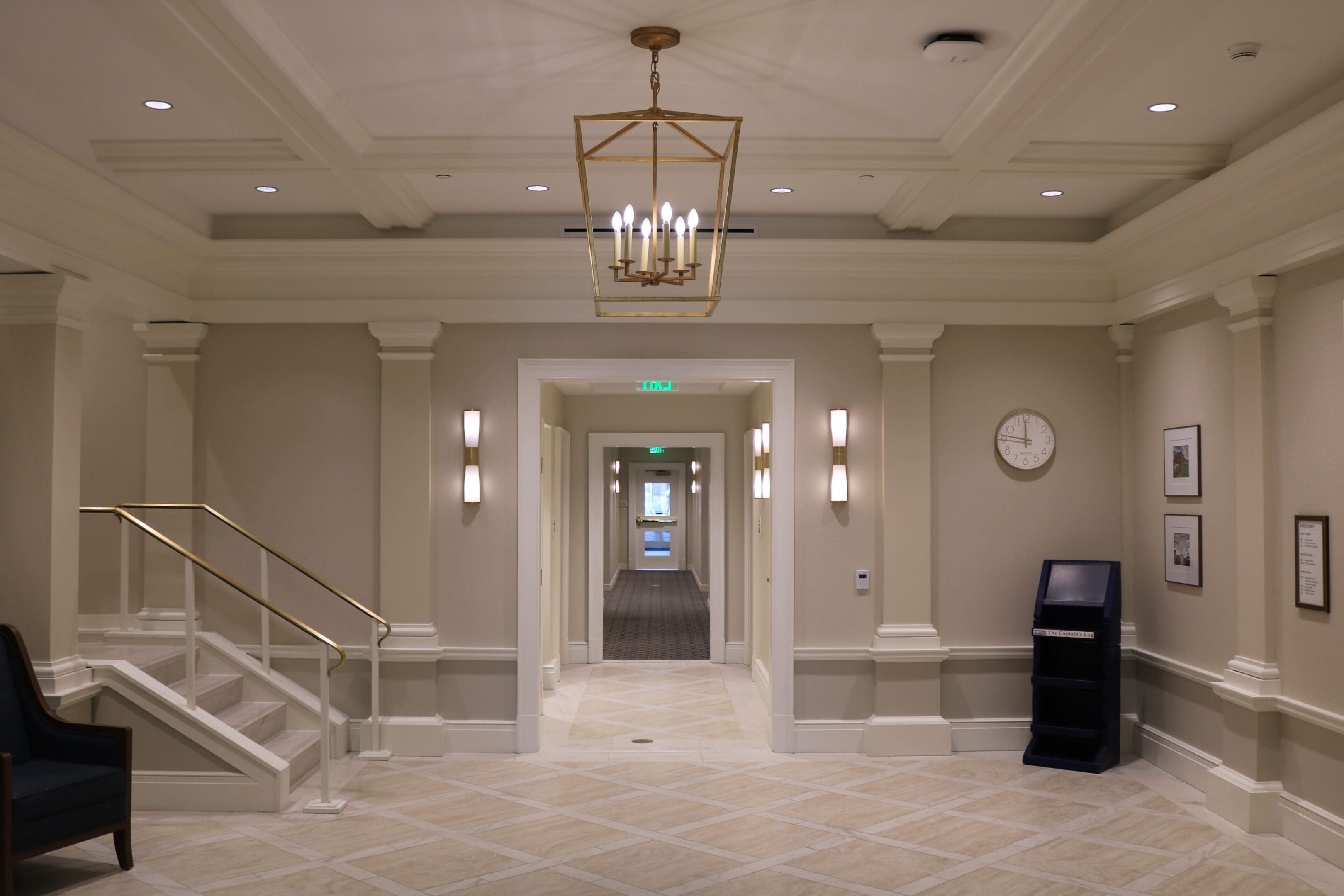 A hallway with a chandelier, staircase on the left, and a clock on the right wall. The corridor is lined with light fixtures and leads to another hallway with a partially open door.