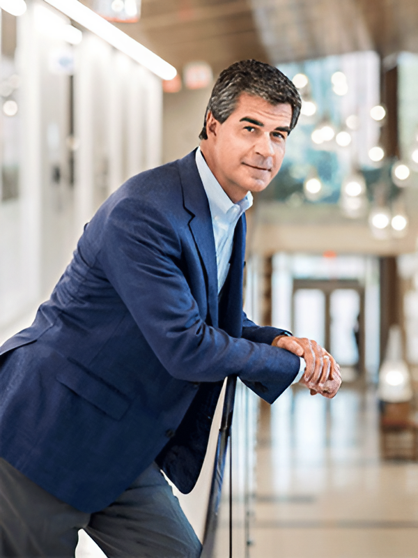 A man in a blue blazer leans on a railing in a brightly lit hallway.