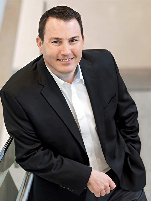 A man in a black suit and white shirt smiles while leaning on a railing indoors.