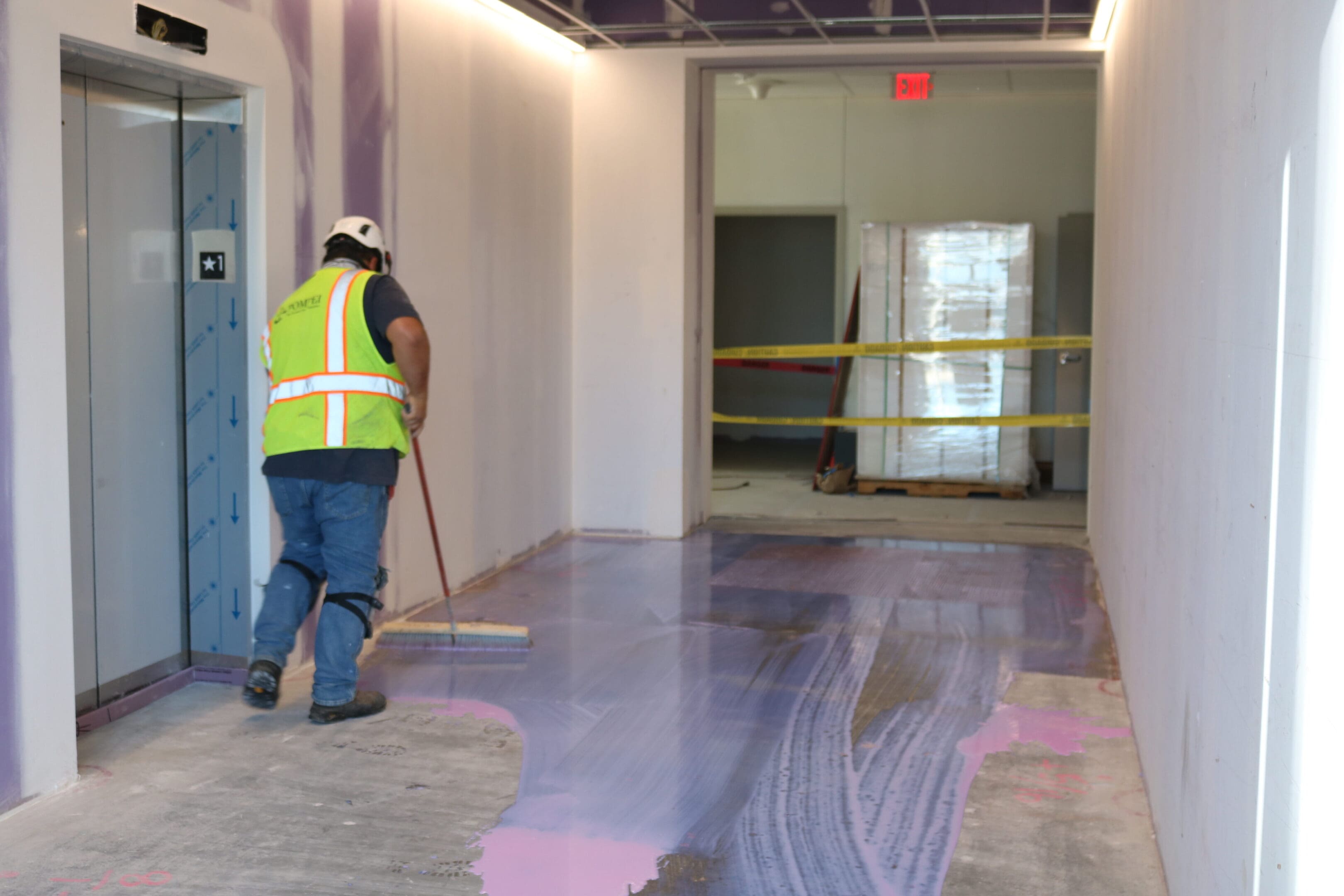 A construction worker in a safety vest sweeps a liquid coating on a corridor floor under renovation. An elevator is on the left and the hallway is taped off in the background.