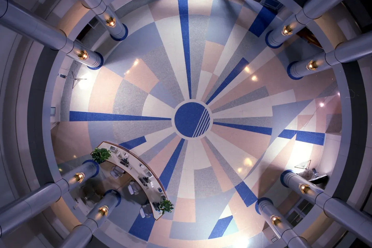 Aerial view of a circular lobby with a starburst pattern on the floor, encircled by columns. A curved reception desk with plants is visible on the lower side.