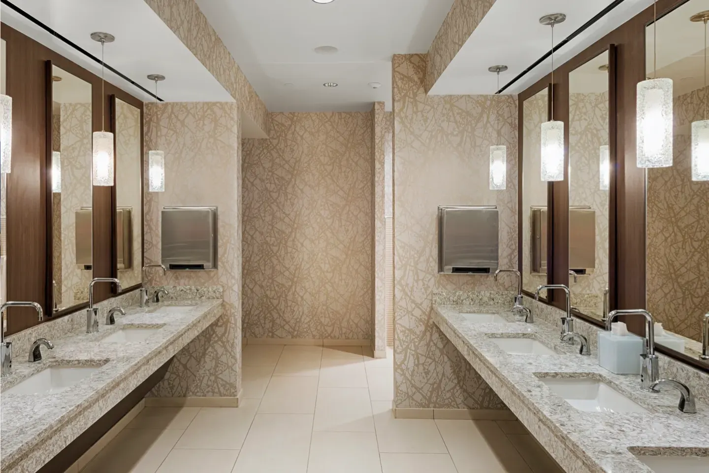 A modern public restroom with two granite-topped sinks, large mirrors, paper towel dispensers, and pendant lights. Cream-colored tiles cover the walls and floor.