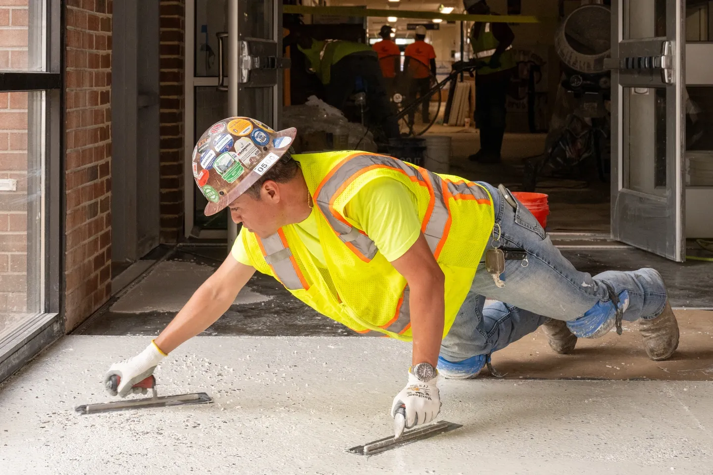 Construction worker smoothing wet concrete with trowels, wearing a hard hat and safety vest inside a building. Several tools and workers in the background.