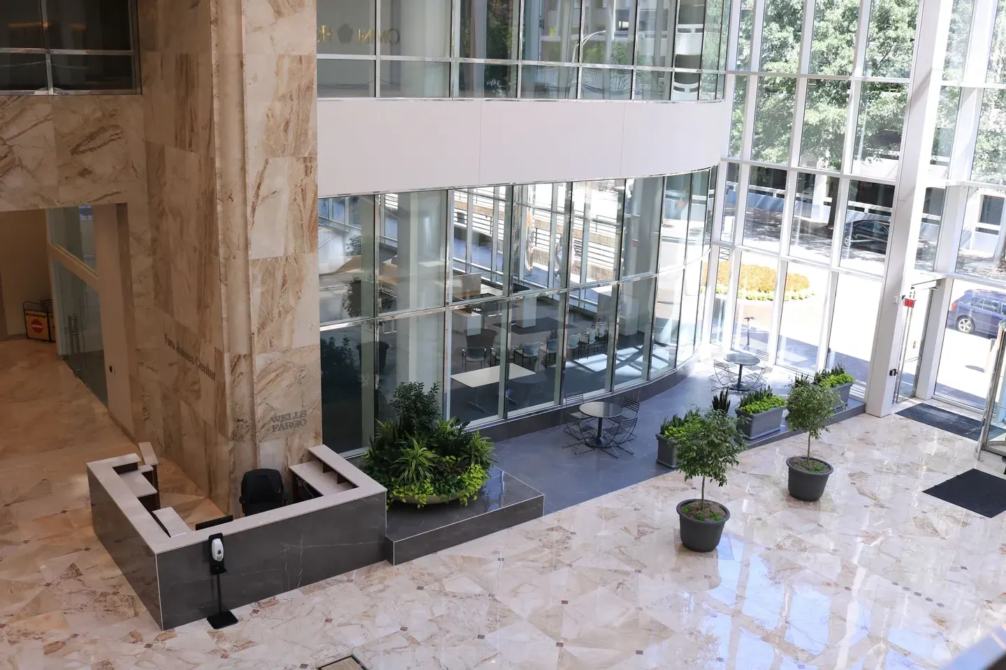 Spacious lobby with marble floors, glass walls, and potted plants. Tables and chairs are placed near the windows.